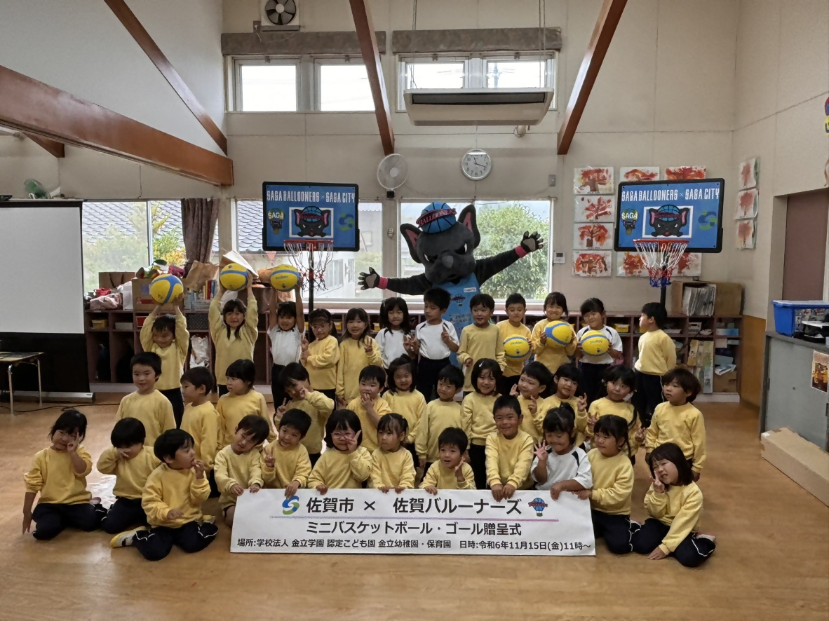 金立幼稚園での贈呈式で、3列に並んだ子供たちの後ろで両手を広げたバルたんが立ち、その両サイドにミニバスケットゴールが置かれている集合写真