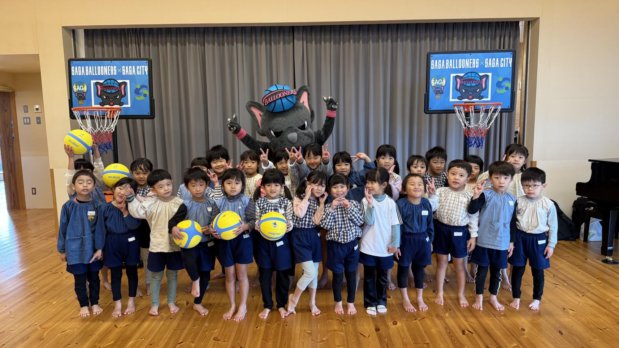 そらいろこども園での贈呈式で、2列に並んだ子供たちの後ろに両手を広げたバルたんが立ち、その両サイドにミニバスケットゴールが置かれている集合写真