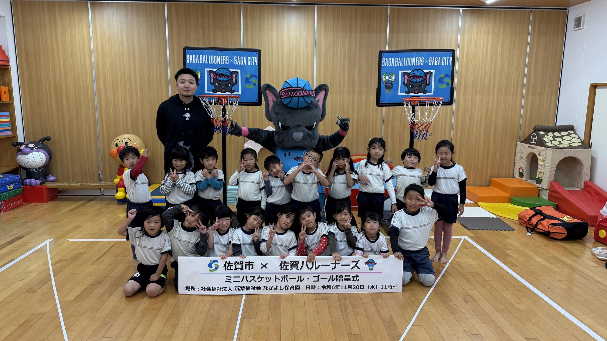 なかよし保育園での贈呈式で、2列に並んだ子供たちの後ろに両手を広げてピースをするバルたんと選手が立ち、両サイドにミニバスケットゴールが置かれている集合写真