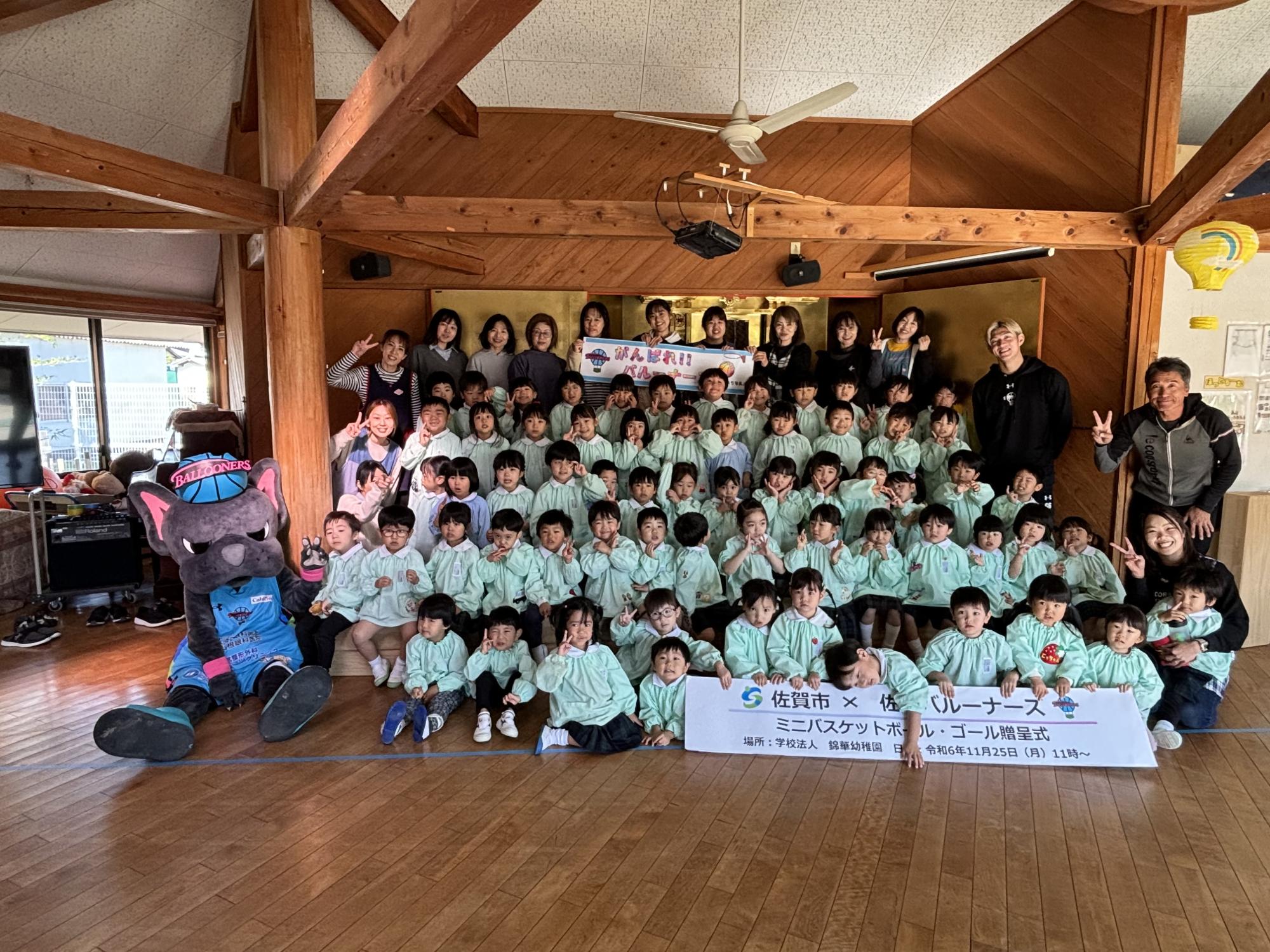 錦華幼稚園での贈呈式で、並んで座るたくさんの園児の左にバルたんが座り、後方に選手や職員の方が並んでいる集合写真