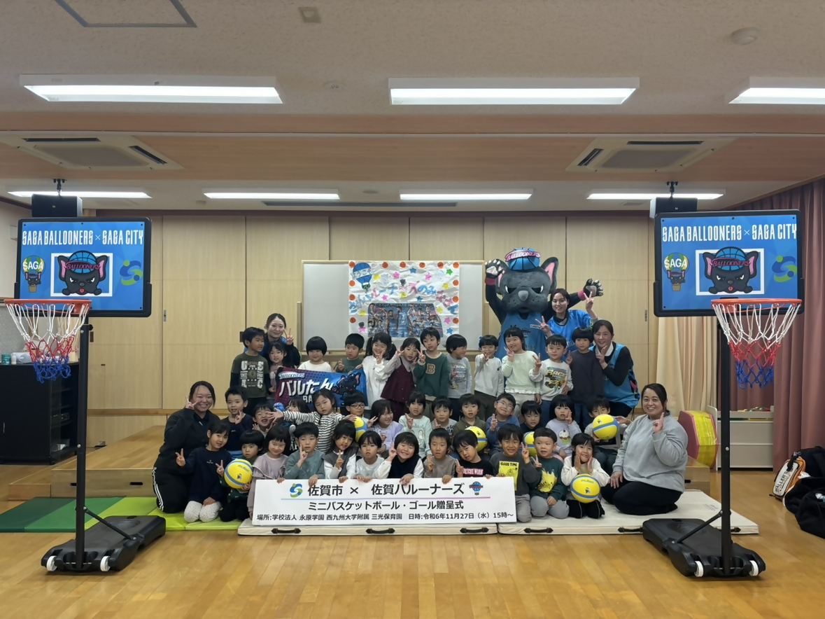 西九大附属三光保育園での贈呈式で、並んで集まった子供たちの後ろにバルたんが立ち、両サイドにミニバスケットゴールが置かれている集合写真