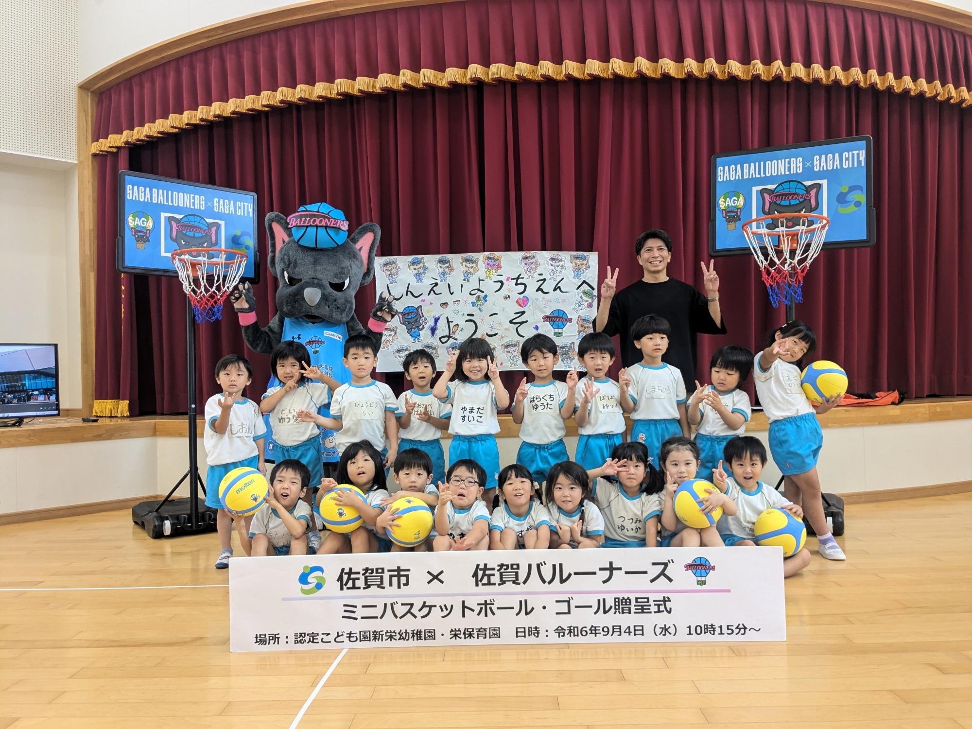 新栄幼稚園での贈呈式で、2列に並んだ子供たちの後ろに佐賀バルーナーズの選手、マスコットキャラクターのバルたんが立ち、両サイドにミニバスケットゴールが置かれている集合写真