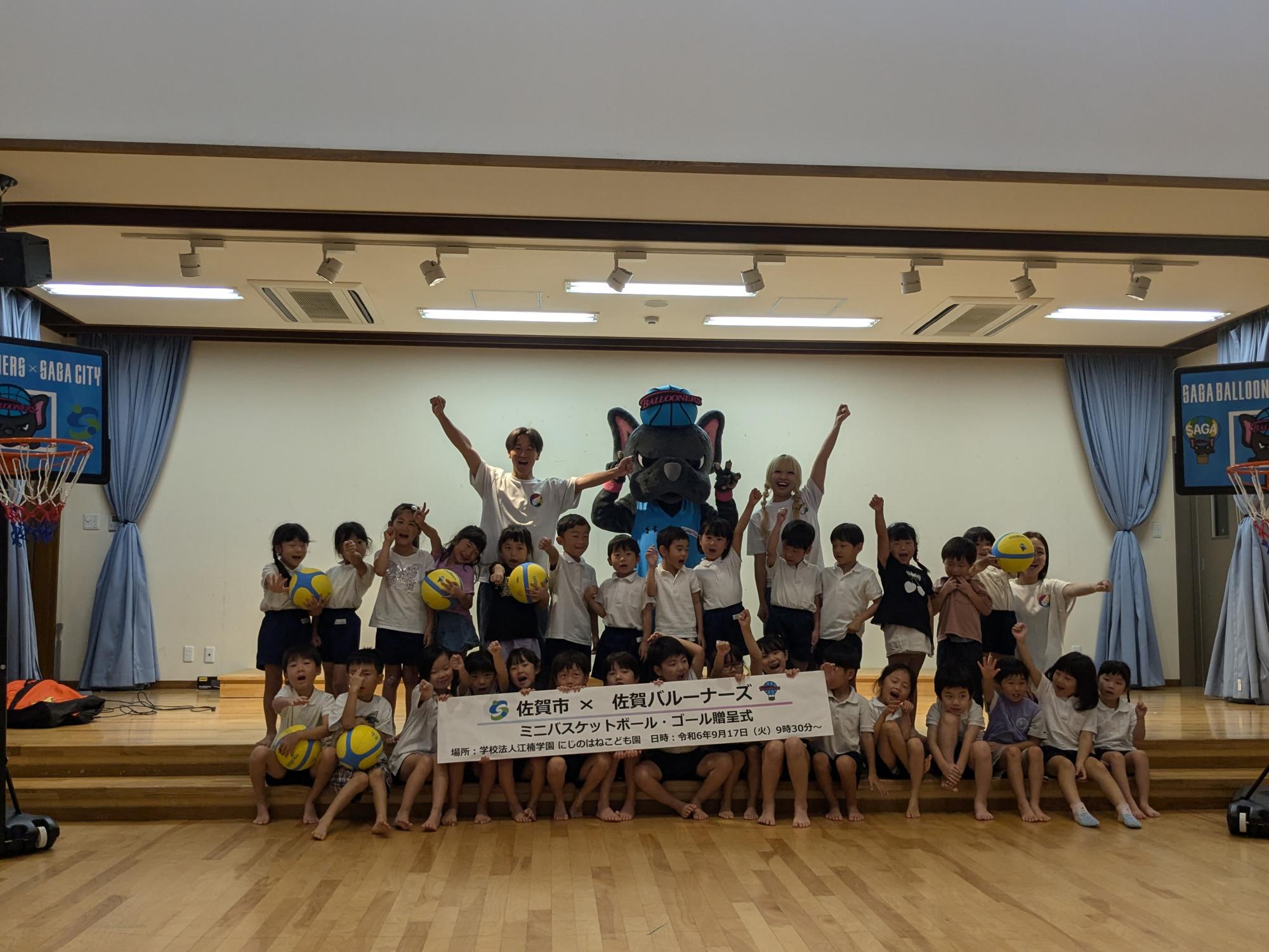 にじのはねこども園での贈呈式で、子供たちが2列に並び、その後ろに職員の方とバルたんが立っている集合写真