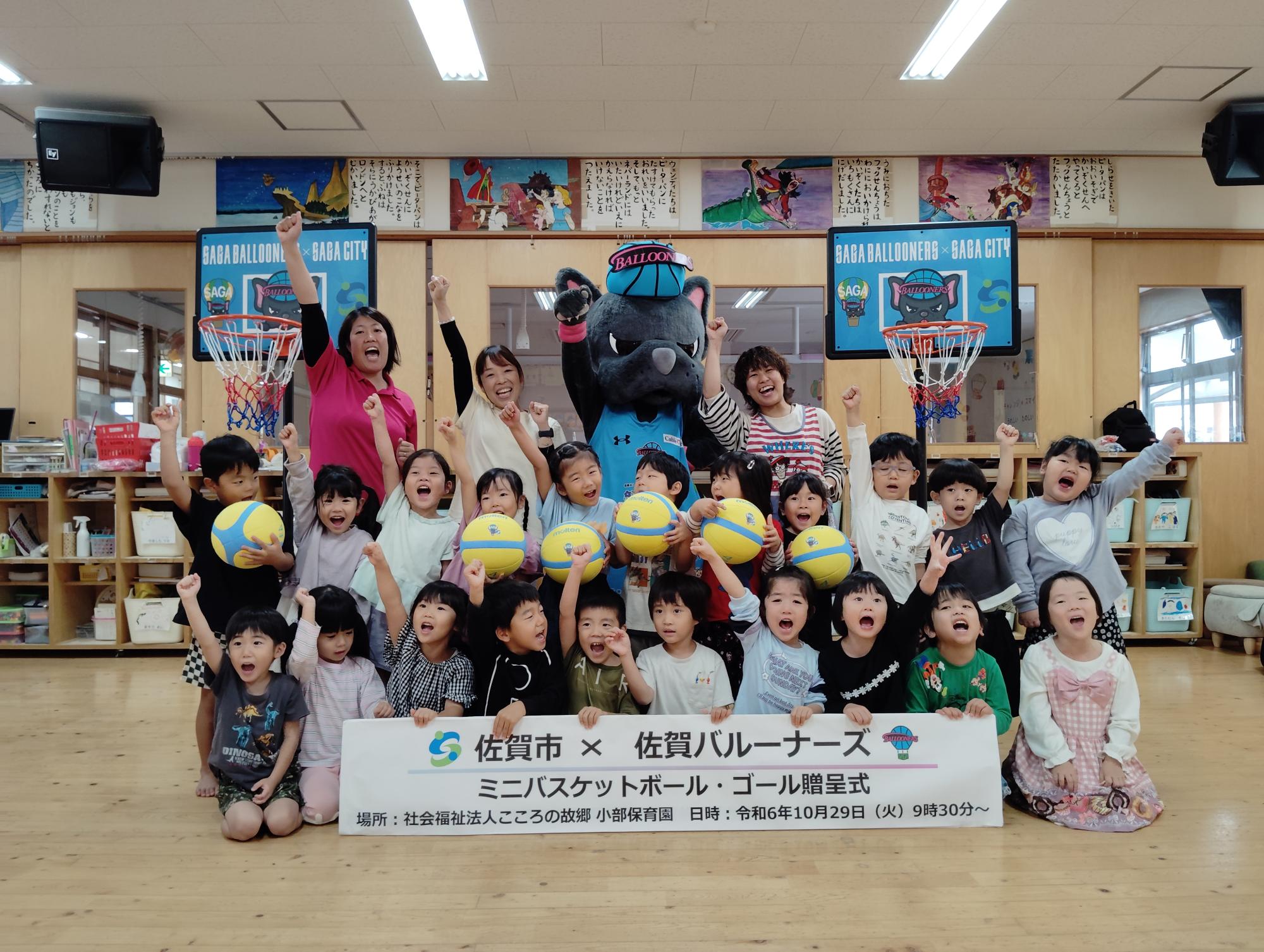 小部保育園での贈呈式で、2列に並び手をあげるポーズをする子供たちと、その後ろに手をあげる職員の方とバルたんが立ち、両サイドにミニバスケットゴールが置かれている集合写真