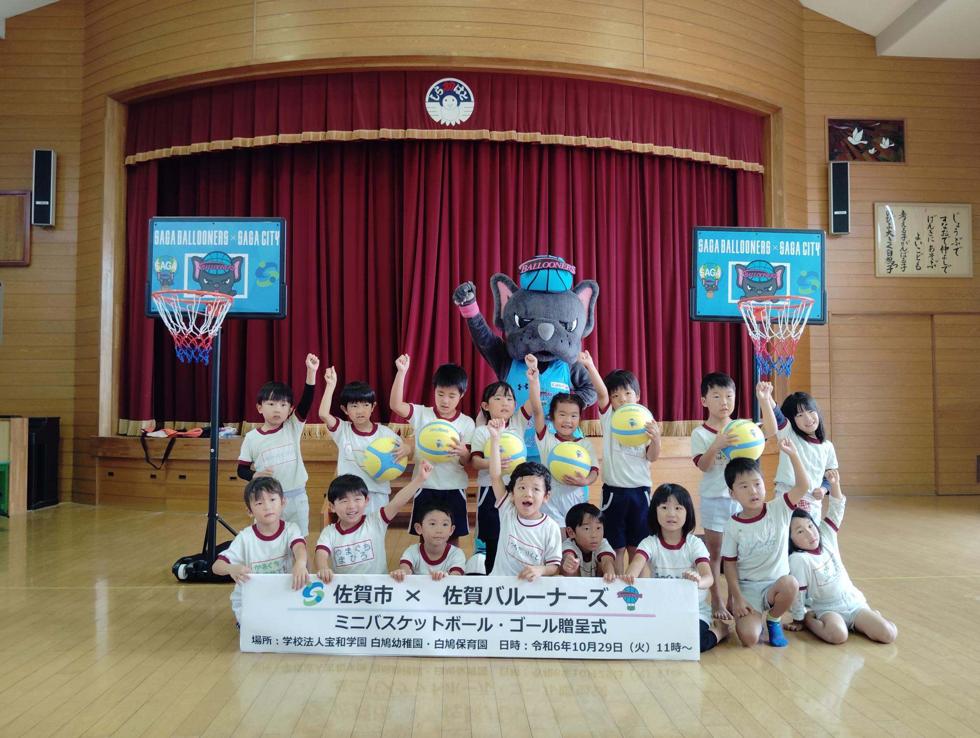 白鳩保育園での贈呈式で、2列に並んだ子供たちが手をあげ、その後ろでバルたんも手をあげて立ち、両サイドにミニバスケットゴールが置かれている集合写真