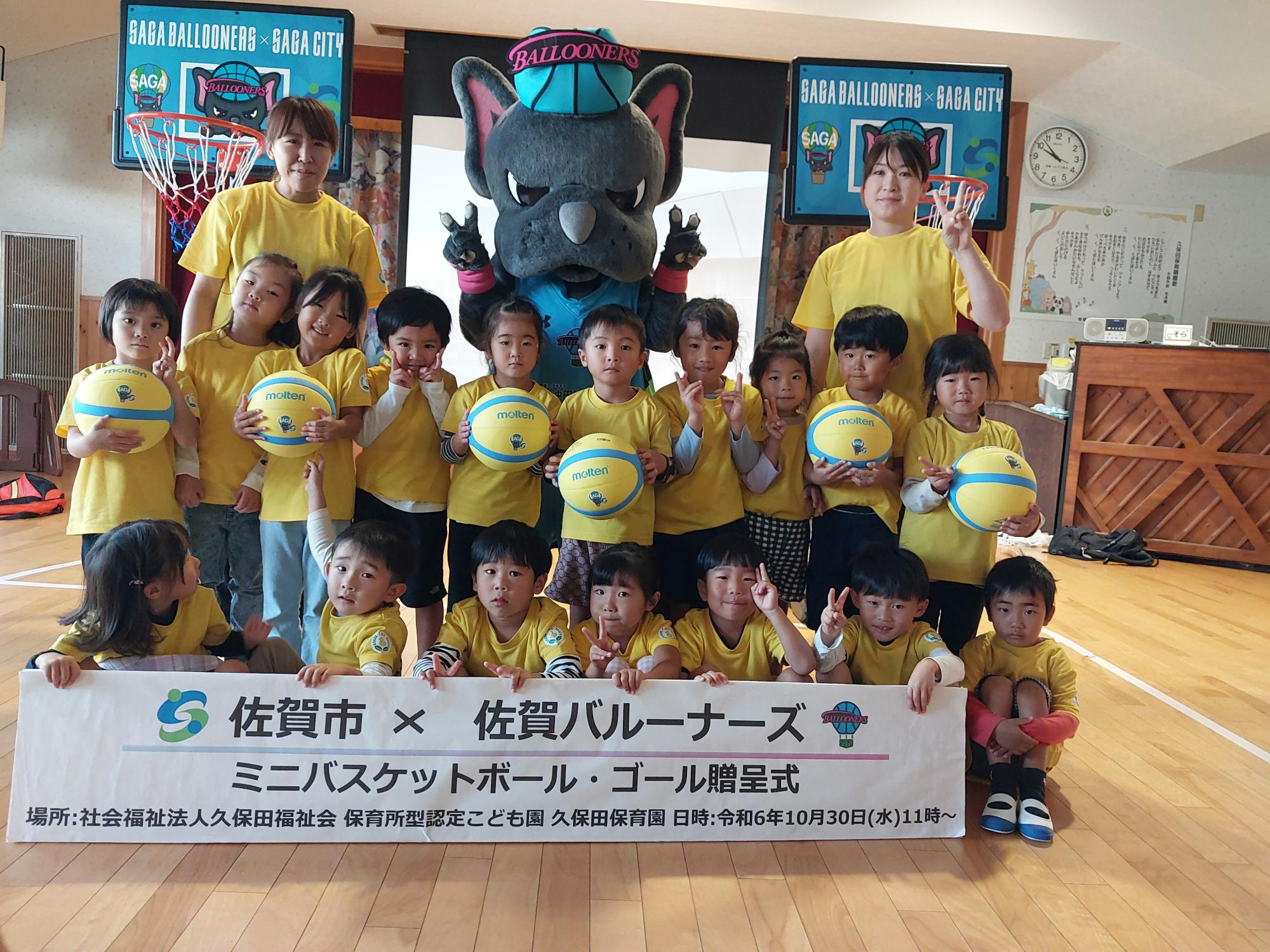 久保田保育園での贈呈式で、2列に並んだ子供たちの後ろに両手でピースをするバルたんと職員の方がならび、その後ろに2つミニバスケットゴールが並んでいる集合写真