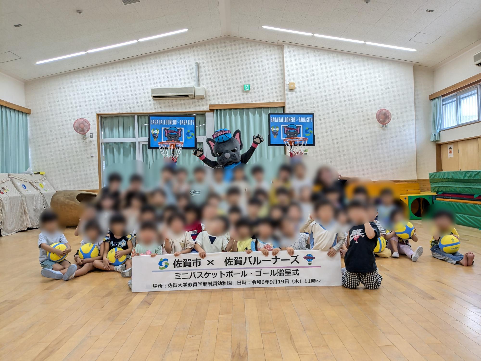 佐賀大学附属幼稚園での贈呈式で、子供たちが並んでいる後ろに、手を広げたバルたんが立ち、その両サイドにミニバスケットゴールが置かれている集合写真