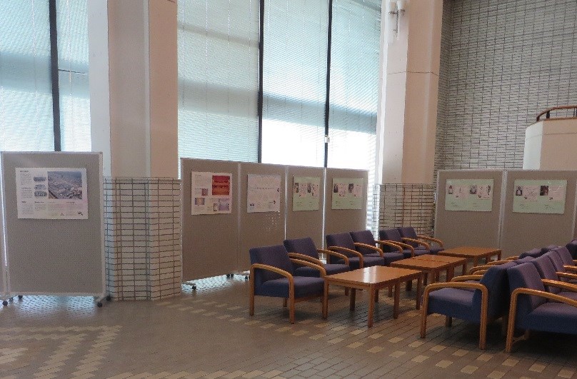 高い天井と大きな窓から光が差し込む明るいロビーに、背もたれ付きの青い椅子と木製テーブルが並ぶ休憩スペースがあり、壁際やパーテーションには地域の歴史や活動を紹介する複数のポスターが展示されている様子を捉えた写真