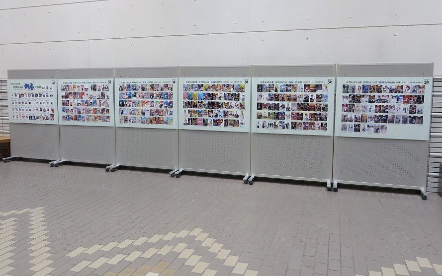 明るい照明に照らされた屋内の展示スペースにて、グレーの可動式パーテーションが六枚横に並び、それぞれの面に過去の公演ポスターや活動の記録と思われる多数の小さな写真や資料が、整然とタイル状に掲示されている様子を捉えた写真