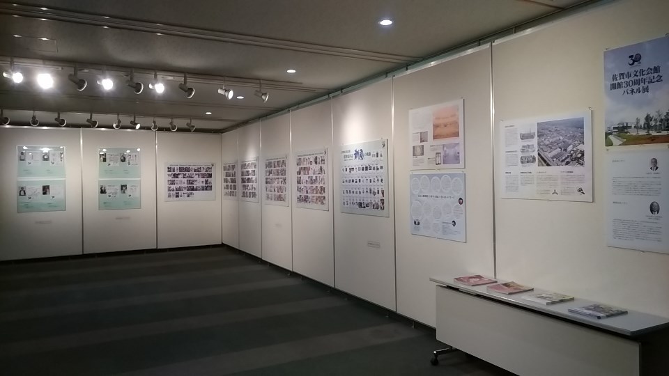 照明を落とした落ち着いた展示室内で、白いパーテーションで区切られた壁面に沿って、佐賀市文化会館の開館30周年を記念した多数の解説パネルや写真資料が整然と並び、手前のテーブルには関連する冊子が置かれている様子を捉えた写真
