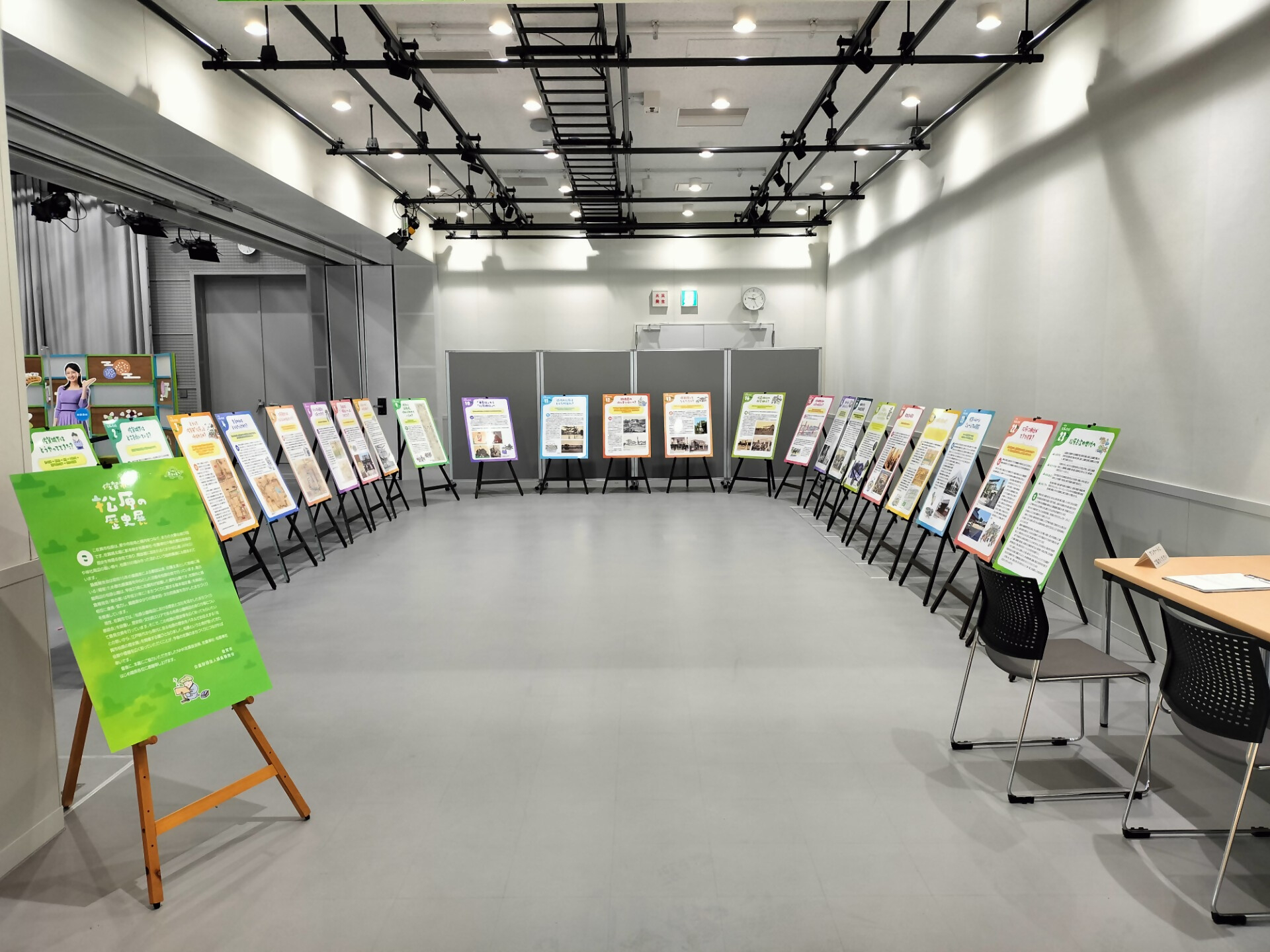 明るい照明に照らされた屋内の展示会場で、壁に沿って半円を描くように二十枚以上の解説パネルがイーゼルに載せられて整然と並び、手前には緑色の大きな紹介ボードや、奥にグレーのパーテーションが配置された佐賀市松原の歴史展会場の展示風景を捉えた写真