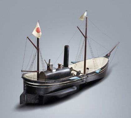 中央に黒い煙突を備えた蒸気機関のボイラーを載せ、船体後方のマストには日の丸が掲げられた、幕末から明治初期の雰囲気を伝える木製の外輪船模型を斜め後ろから捉えた写真