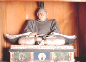 木製の厨子の中に安置された、黒い束帯姿に冠を戴き、両手を膝の上で組んで座る、彩色が施された台座に乗った鍋島忠直坐像の木造彫刻を正面から捉えた写真