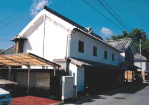 真っ白な漆喰の壁と重厚な瓦屋根を持つ土蔵造りの建物が道路沿いに並び、手前の駐車場には白い車が停まっている様子を捉えた写真