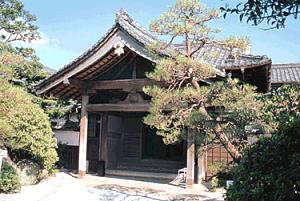立派な瓦屋根と太い木製の柱が特徴的な日本家屋の玄関先を、手前に植えられた松の木や周囲の豊かな緑とともに捉えた旧古賀家の外観写真