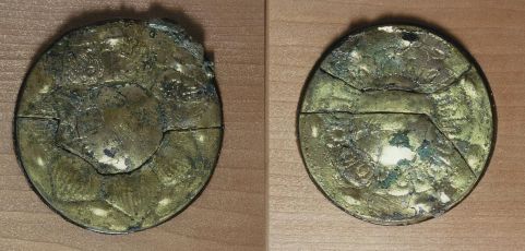 経年による摩耗や亀裂が見られる金色の円形金具が上下に二点並び、蓮華文のような装飾が浮き彫りにされた表面の質感や、一部に緑青が残る古色蒼然とした石塚一号墳出土遺物の写真