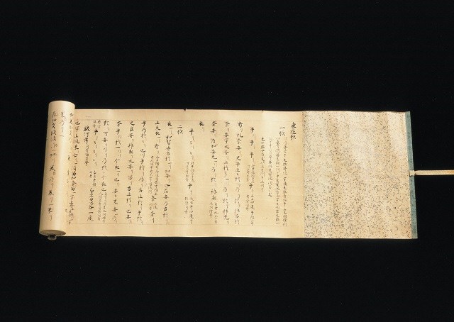 漆黒の背景の中で左側に芯を残して横長に広げられた1巻の巻物であり、紙面には墨で細かく丁寧に記された古文書の本文が並び、右端には銀色の細かな斑点模様が散りばめられた上品な装飾が施されている、保存状態の良さと洗練された美しさを併せ持つ紙本墨書東遊歌神楽歌の写真