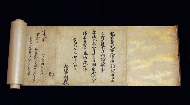黒い背景を背に、左側に芯の部分が見える状態で横長に広げられた1巻の巻物であり、紙面には墨で力強く書かれた漢字の古文書と独特な署名が記され、右端には金箔を散らしたような華やかな装飾が施された、美術的価値と歴史の深さを感じさせる松浦山代家文書の写真