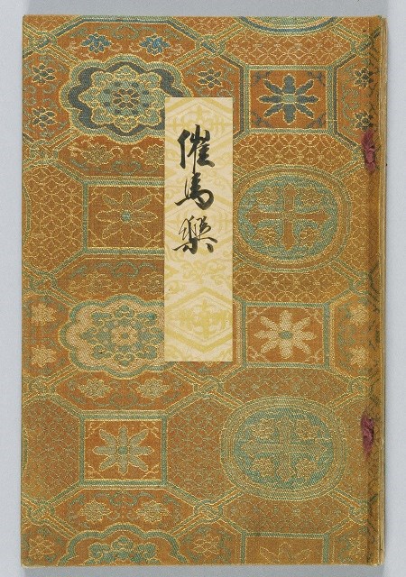 金糸や色糸を用いて亀甲文や花菱文といった緻密な幾何学模様が織り込まれた、格調高い装飾文様の布張りの表紙に、中央に「催馬楽」と墨で力強く題字が書かれた白い題箋が貼られた、日本の伝統的な装丁の美しさを伝える古い催馬楽譜の写真