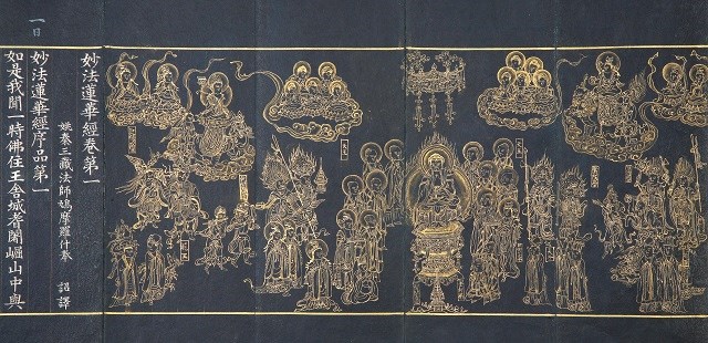 金地に細密な線で多数の仏像や宝相華、雲上の群像が左右に広がり、左端に縦書きの金文字が配された荘厳な仏教画面を示す写真