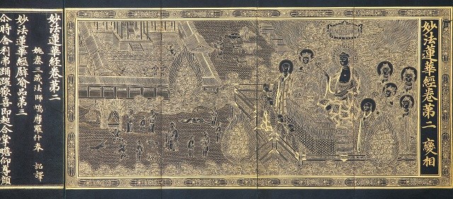 金地に黒線で細密に建物や樹木、人物群像、中央の座像などが描かれ、左右に縦書きの金文字が配された荘厳な仏教画面を写した写真