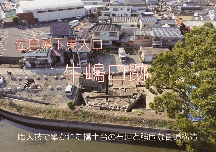 川沿いで行われている発掘調査現場を上空から俯瞰で捉えた、写真の中央に赤縁の白い文字で「佐賀城下東入口 牛嶋口跡」のタイトルが、下部に白い文字で「職人技で築かれた橋土台の石垣と強固な街道構造」と説明文が配置された現場写真