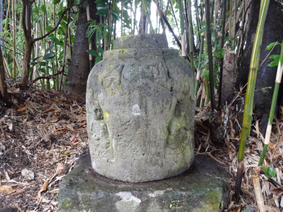 竹林の根元にひっそりと佇む、側面に小さな仏坐像の浮き彫りと無数の漢字が刻まれた円筒形の古い石造物の写真