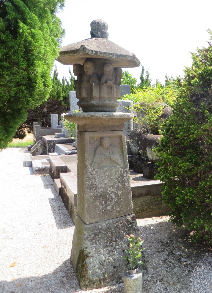 墓地の一角で、火袋の部分に複数の小さな仏像が浮き彫りにされ、中台の下の竿部分にも仏坐像が彫り込まれた特徴的な石灯籠が、周囲の木々や墓石に囲まれいる写真