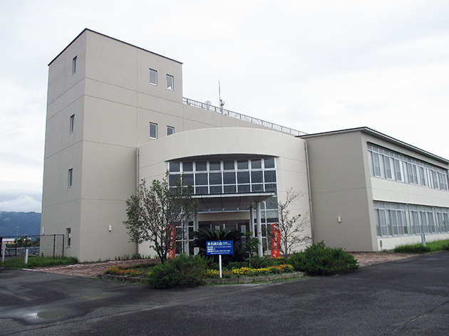 ベージュの外壁を持つ3階建ての公共施設のような建物が立っており、エントランス部分には円弧を描くデザインの窓と青い案内看板、そして植栽や赤い幟が配置されている東名縄文館外観写真