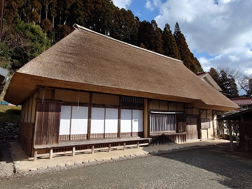 青空の下で山林を背に立つ、美しく整えられた大きな茅葺き屋根が特徴的な木造の古民家であり、白い障子がはめ込まれた縁側や土間へと続く入り口が見える、吉村家住宅の写真