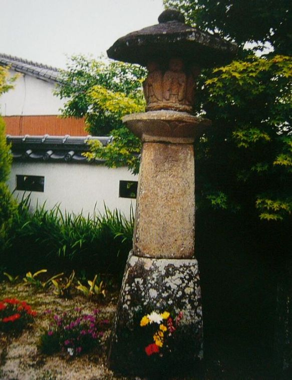 白壁の建物と青々とした樹木を背景に、火袋の部分に複数の仏像が浮き彫りにされた古びた石灯籠が立ち、その足元には黄色や赤色の鮮やかな供花が供えられた久留間六地蔵の写真