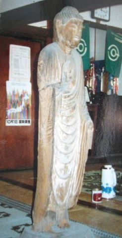 室内に安置された木彫りの仏像が流れる衣文の彫りや端正な立ち姿を示し、周囲の道具類や掲示物に囲まれながら静かな存在感を放つ写真