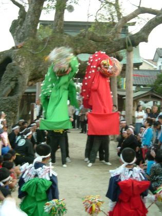神社の境内に立つ巨木を背景に、鮮やかな緑色と赤色の長い布を垂らした二体の獅子頭が、大勢の見物客が見守る中で天高く掲げられ勇壮に舞っている様子の写真