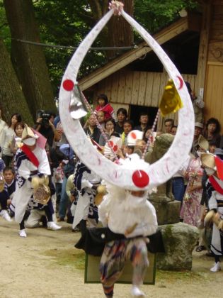 神社の境内で、大きな円形の白い飾りを頭に戴いた踊り手が激しく舞い、その周囲を鉦を鳴らす演者や大勢の見物客が囲む行事の様子の写真