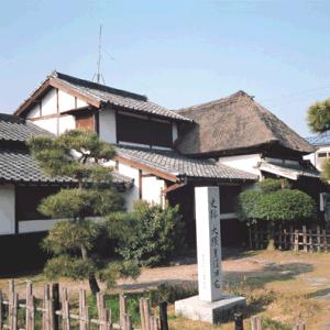 茅葺き屋根と瓦屋根が混在した伝統的な日本家屋の前に「史跡 大隈重信旧宅」と刻まれた白い石碑が建ち、手前には松の木と竹垣が配置された大隈重信旧宅の外観写真