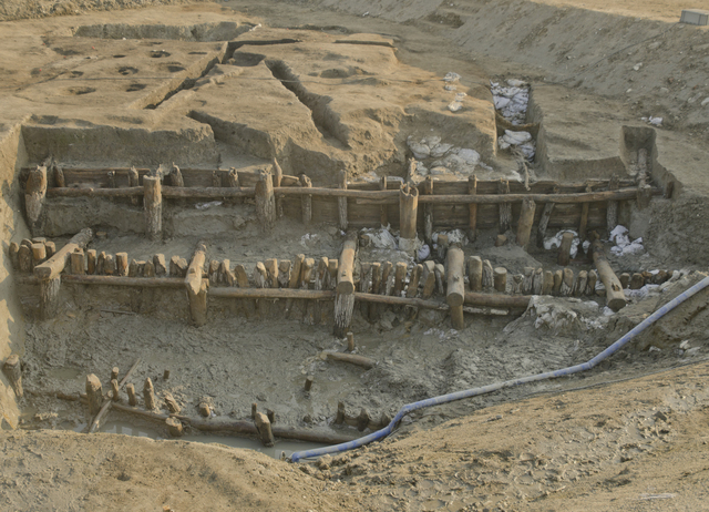 広大な発掘調査現場において、湿った粘土質の地面に縄文時代のものと思われる木柱や横木が複雑に組み合わされ、当時の高度な土木技術を物語るような大規模な遺構が剥き出しの状態で保存されている遺跡の写真