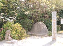 陽光が差し込む明るい屋外で、手前に「鍋島直茂誕生地」と刻まれた白い石柱が立ち、その奥にこんもりとした丸い岩のような石碑と、小さな仏像のような石造物が緑豊かな樹木を背景に並んでいる、鍋島直茂誕生地の写真
