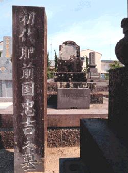 晴天の下、墓地に建つ複数の古い墓石の中で、手前に「初代肥前国忠吉之墓」という文字が深く刻まれた大きな石柱が垂直に立ち、その奥に歴史を感じさせる重厚な石造りの墓が鎮座している様子を捉えた、初代肥前国忠吉の墓地の写真