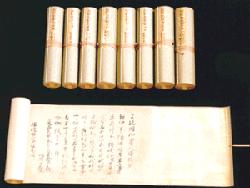 黒い背景の上に、綺麗に巻かれた状態で並ぶ8本の巻物と、その手前で横長に広げられた1本の巻物が置かれており、紙面には墨でさらりと流れるような筆致の古文書が記された、歴史の重みと保存状態の良さを感じさせる深堀家文書の写真