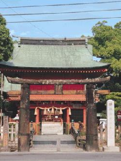 青空の下に佇む朱塗りの鮮やかな社殿を背景に、年月を経て黒ずんだ石造りの鳥居が力強く建っており、注連縄が飾られた拝殿へと続く参道の脇には白い石碑が並ぶ、歴史ある神社の厳かな雰囲気を感じさせる与賀神社三の鳥居の写真