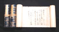 紺色の表紙に金色の文様が施された二本の巻物が紐で括られて左側に並び、右側には白い紙面に墨で流麗な文字が書き連ねられた別の巻物が水平に広げられている深江家文書の写真