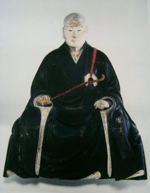 白頭の僧侶が黒い法衣を纏い、赤い紐のついた細長い如意を右手に携えて静かに坐す姿を再現した彫像であり、穏やかな表情と威厳に満ちた佇まいが白背景の中に際立つ、無著妙融像を正面から捉えた写真