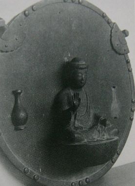 円形の金属板に仏像が浮き彫りにされた懸仏であり、中央で結跏趺坐する仏の傍らに小さな花瓶が配され、縁には等間隔に鋲が並ぶとともに上部に吊り下げ用の金具が備わった懸仏を斜めから捉えた写真