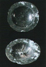 黒っぽい色味の金属で作られた円盤状の遺物が上下に二点並んでおり、上側はなだらかに盛り上がった半球状の表面を、下側は中央に穴が開いた窪みのある裏面を見せている銅釦の写真