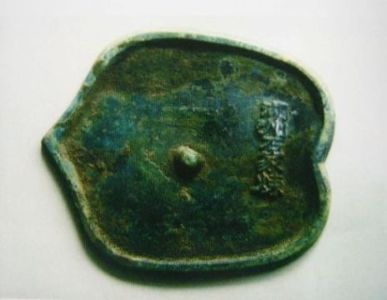 緑青が全体に付着した青銅製の平たい遺物であり、ハート型や葉のような独特の輪郭を持ち、表面の右側には微細な文字のような意匠が浮き彫りにされている、長い年月を経て土中から発見された一本木遺跡出土遺物を上から捉えた写真