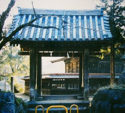 青瓦の切妻屋根を戴く重厚な門が、細い注連縄を下げて静かに佇んでおり、門越しには奥行きのある境内の社殿や周囲を囲む板塀がのぞく、与止日女神社の西門を日差しの中で捉えた写真