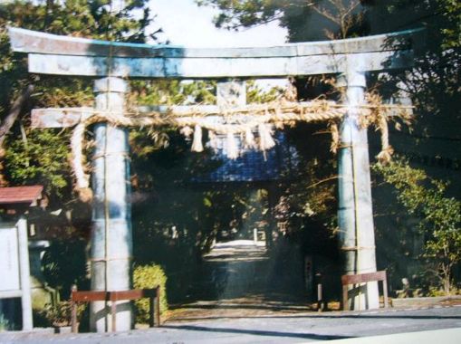 青銅特有の緑青が吹いた石造りのような質感を持つ鳥居が立ち、太い注連縄が掛けられたその先には木々に囲まれた社殿へと続く参道が伸びている、神聖な銅造明神鳥居を日差しの中で穏やかに捉えた写真