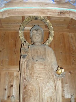 木彫りの素朴な質感を残した薬師如来立像が、右手を施無畏印に結び左手に金色の薬壺を載せて木造の厨子内に佇む、後光を表す円形の光背を背負い静かに人々の平癒を願うような慈悲深い姿を正面から捉えた写真