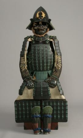 黒い兜に金色の前立てを戴き、緑色の威し糸で綴じられた精巧な大鎧が、重厚な面頬や籠手、脛当と共に飾られている、青漆塗萌黄糸威二枚胴具足の写真