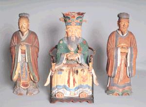 明るいグレーの背景に、鮮やかな色彩が残る三体の木造坐像が並び、中央には豪華な冠を被り緑や黄色の装束を纏った威厳ある人物が座り、その両脇には控えめな褐色の衣を着た二体の人物が直立する鬼丸聖堂の聖像三体の写真